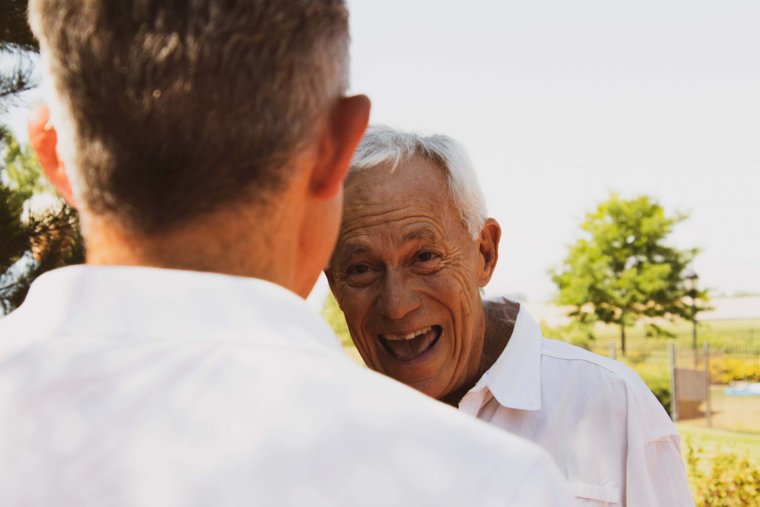 A smiling elderly man in a white shirt faces another person, partially seen from behind, in what appears to be an outdoor setting with trees in the background.