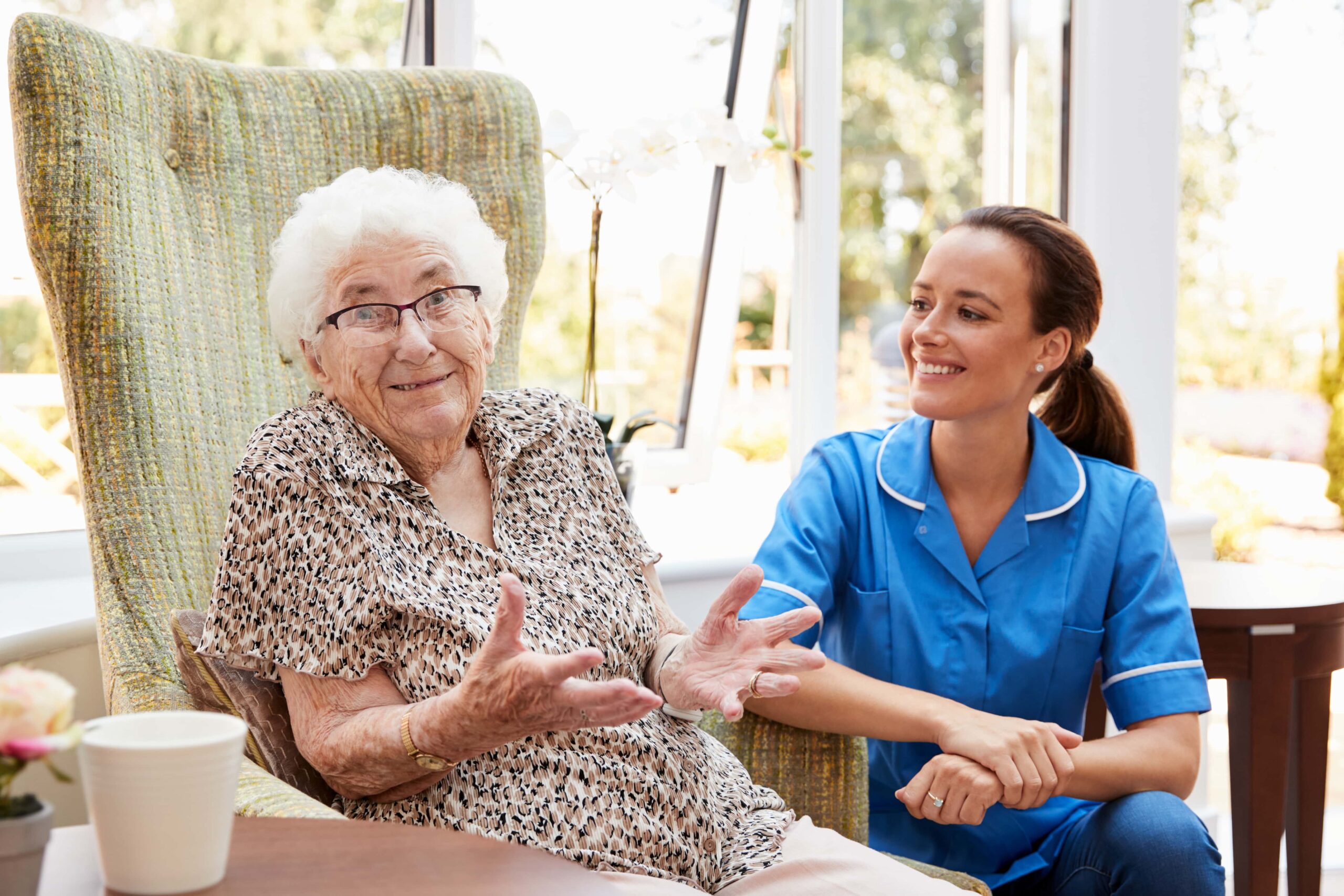 An elderly woman seated in a chair gestures with her hands while a nurse in a blue uniform kneels next to her, smiling in a bright, sunlit room.