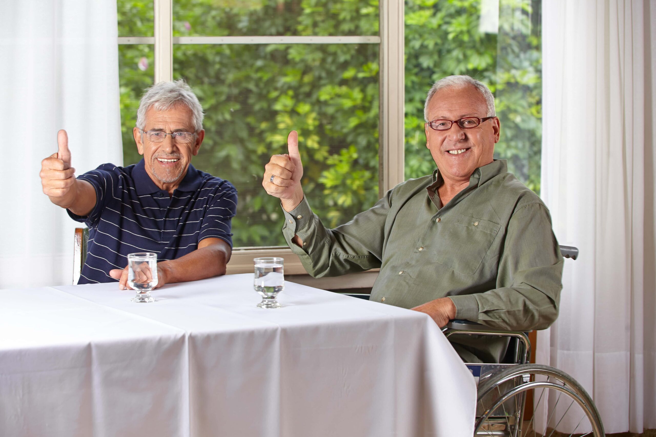 Two elderly men, one sitting in a wheelchair, are indoors at a table with white tablecloths, giving thumbs up and smiling, with a window and green foliage in the background.