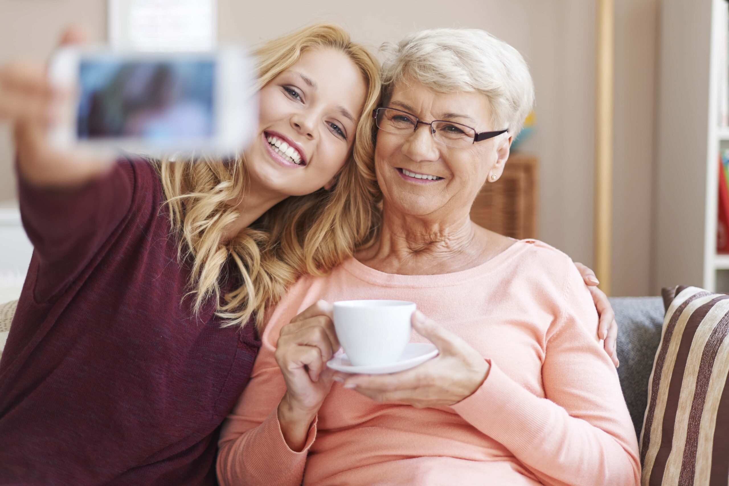 A young woman and an older woman sitting on a couch, smiling and taking a selfie together. The older woman is holding a white cup.