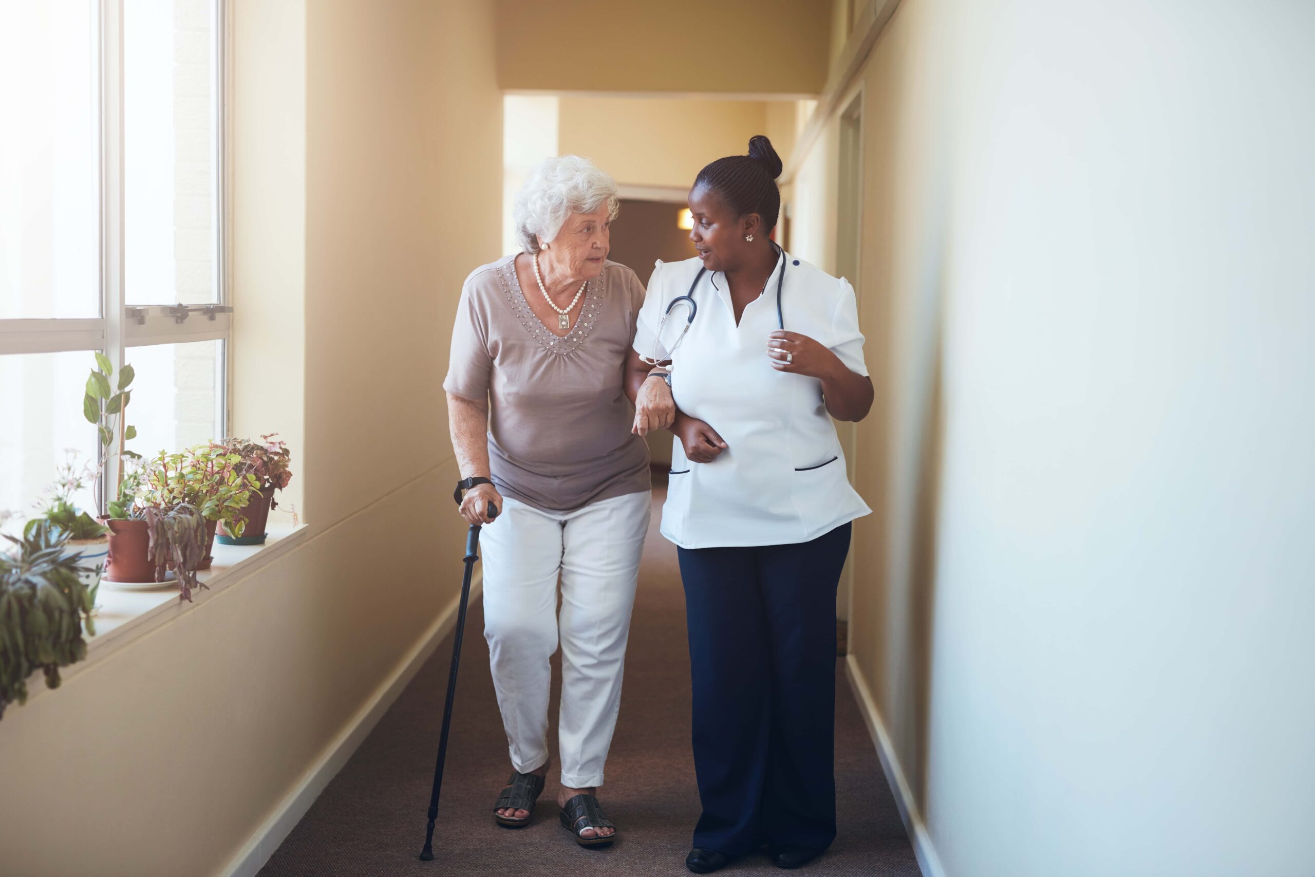 A nurse assists an elderly woman with a cane as they walk down a hallway decorated with potted plants.