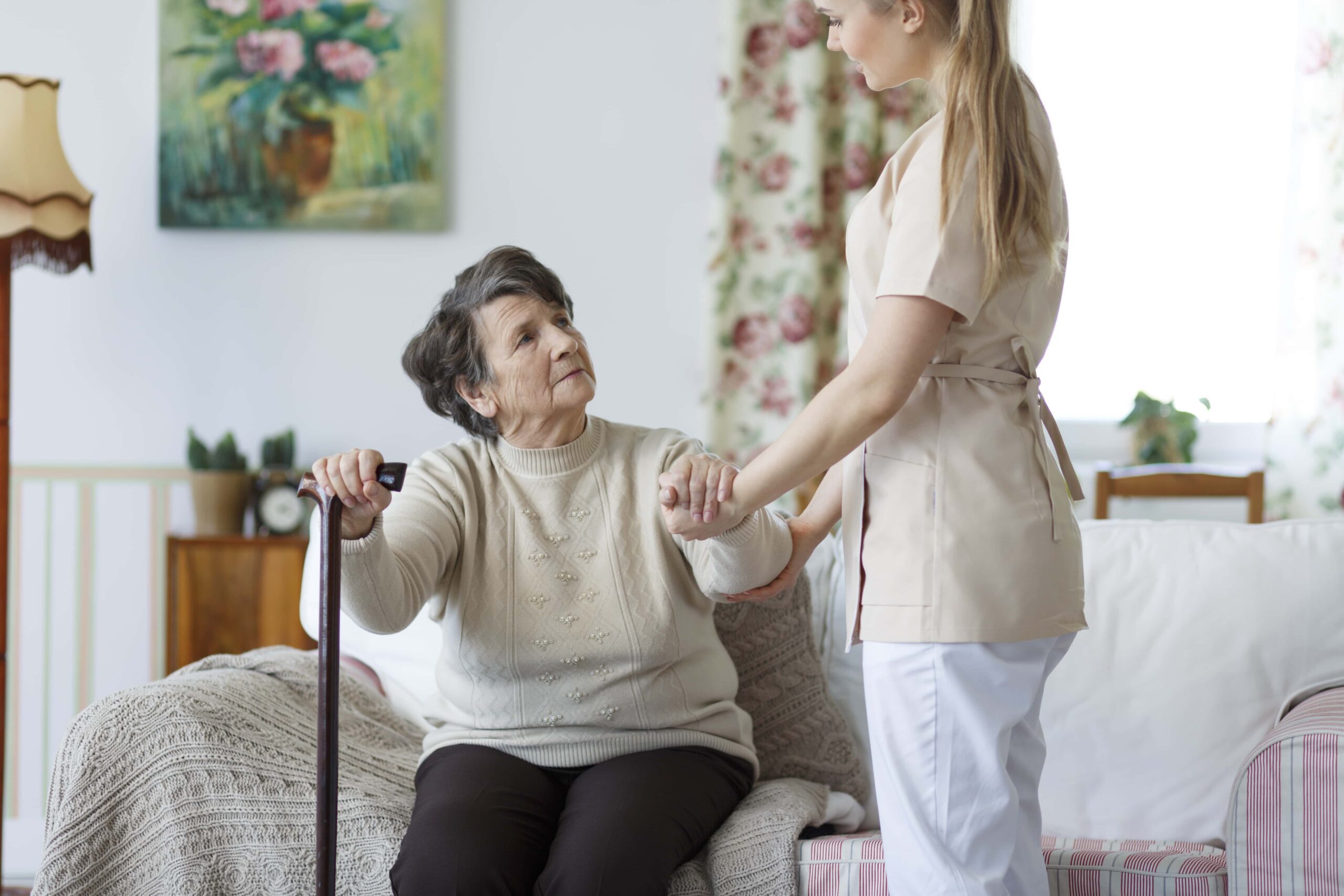 A caregiver assists an elderly woman with a cane in a cozy living room decorated with floral elements.