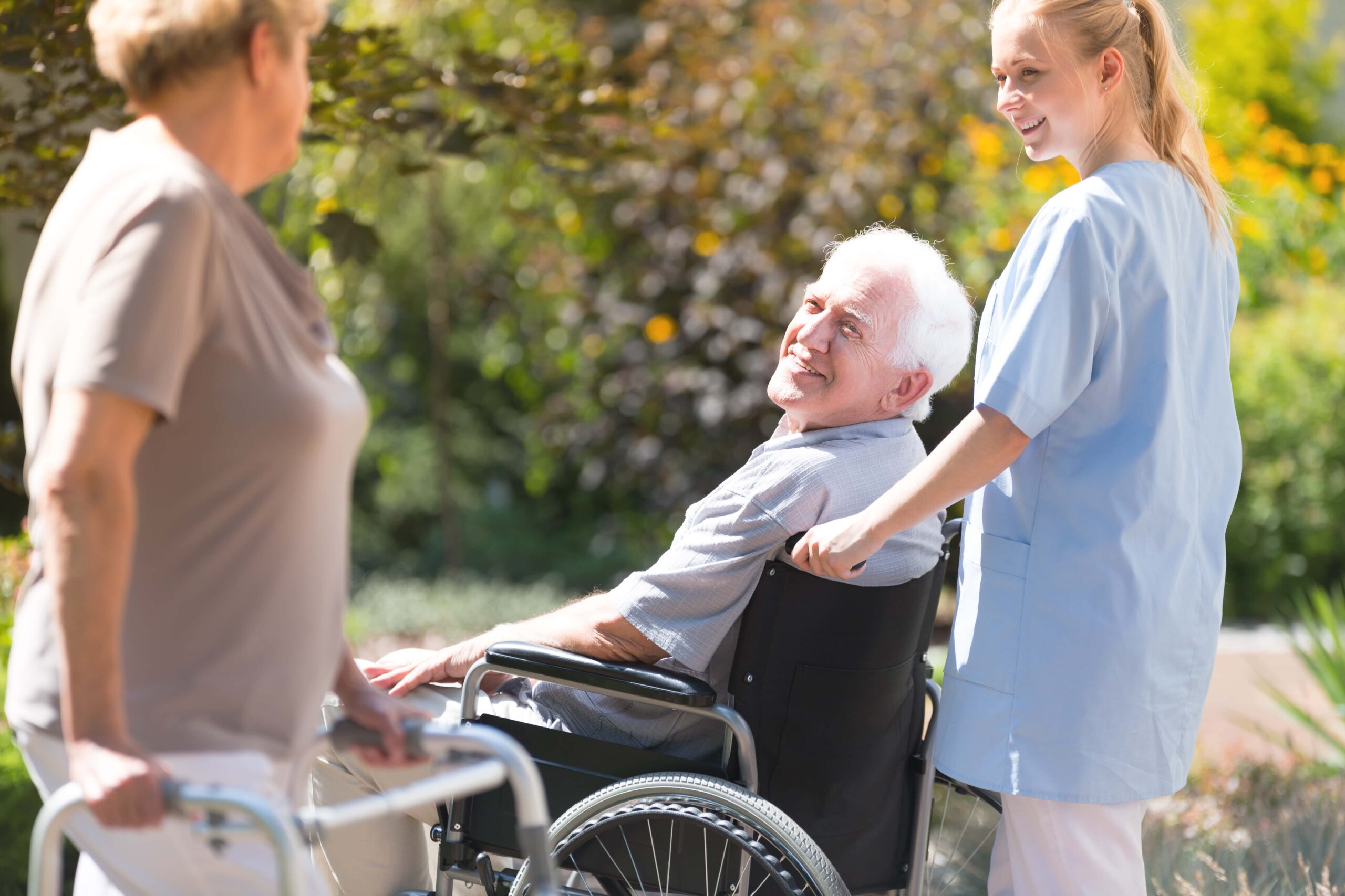 A caregiver in blue scrubs pushes an elderly man in a wheelchair outdoors while an elderly woman with a walker stands nearby.