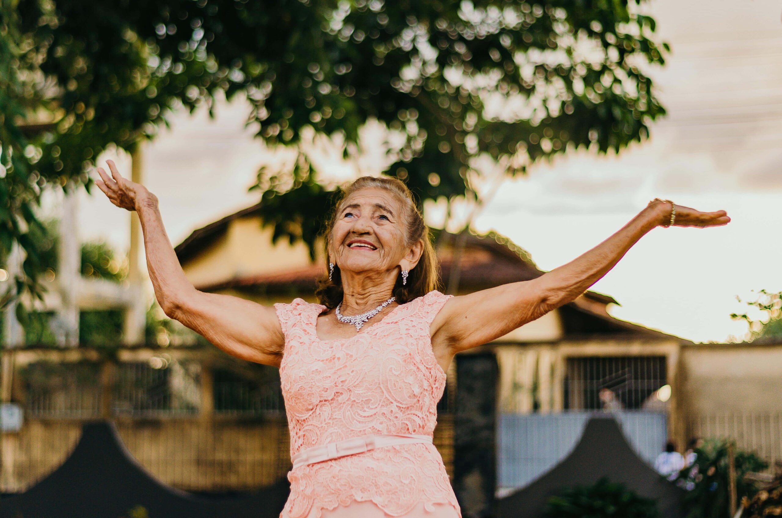 An elderly woman wearing a pink lace dress and necklace raises her arms joyfully outdoors, with trees and buildings in the background.
